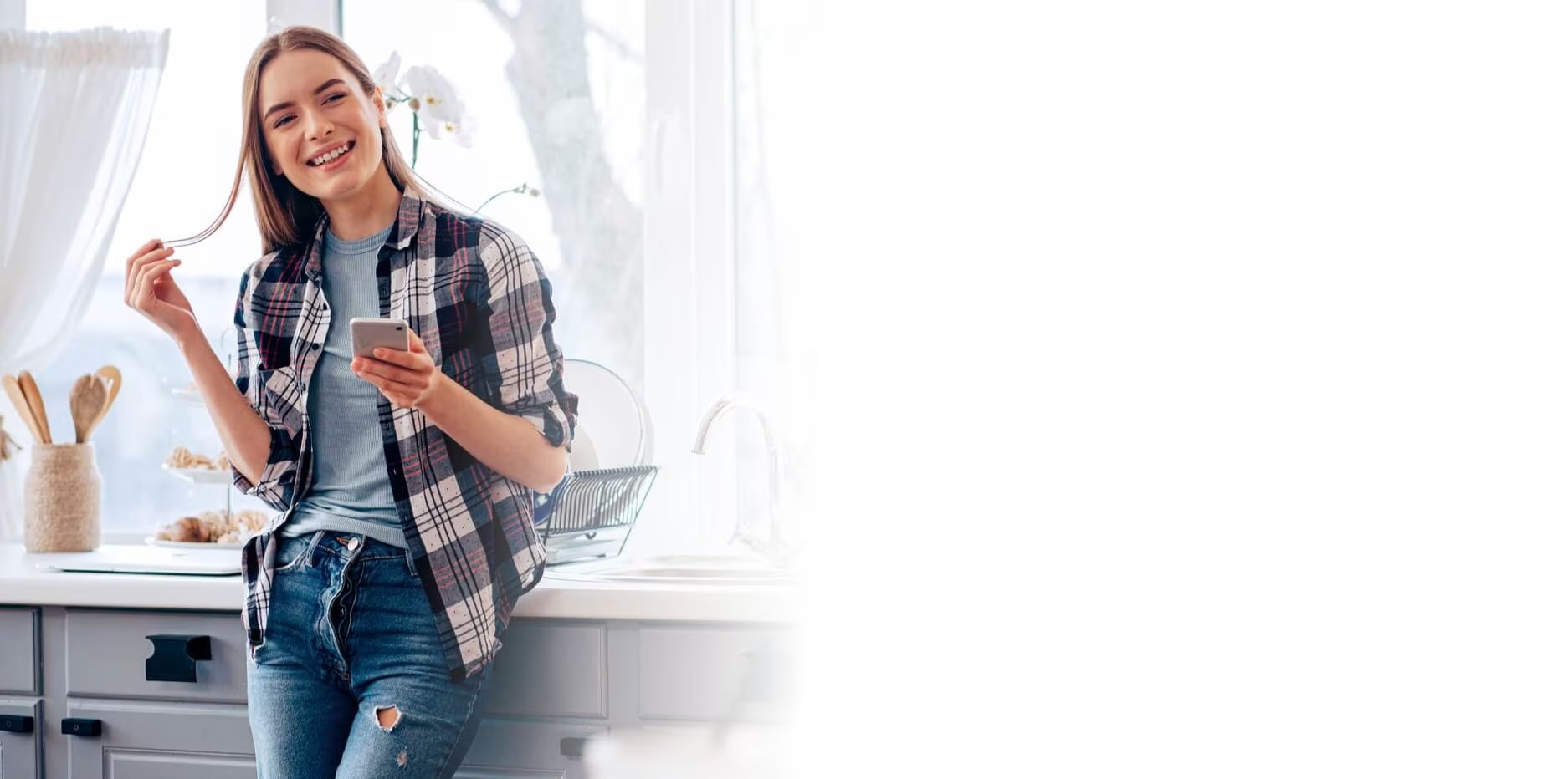 Smiling young woman in a plaid shirt and jeans holding a smartphone and playing with her hair in a bright kitchen with utensils and dried flowers on the counter