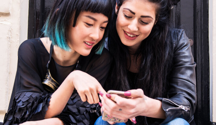 Two young women sitting together in front of a black door, smiling and looking at a smartphone, one with short black hair dyed turquoise at the ends and wearing a black top, the other with long black hair styled in two buns, wearing a black leather jacket and blue jeans.
