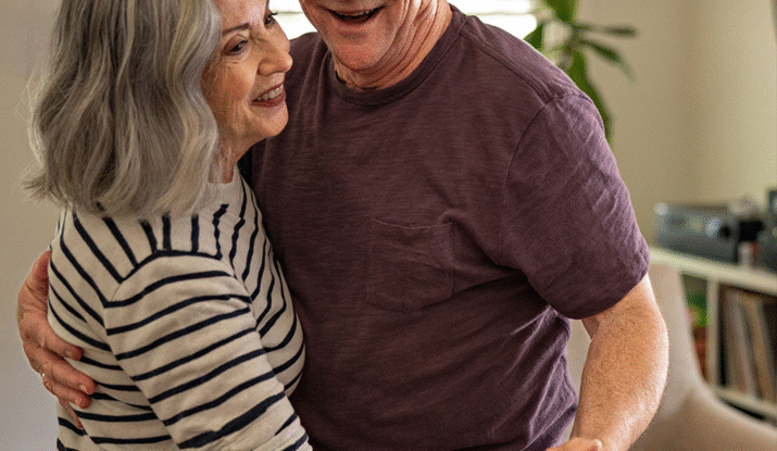 Older couple smiling and dancing closely indoors, with the woman wearing a striped shirt and the man in a purple shirt and glasses.