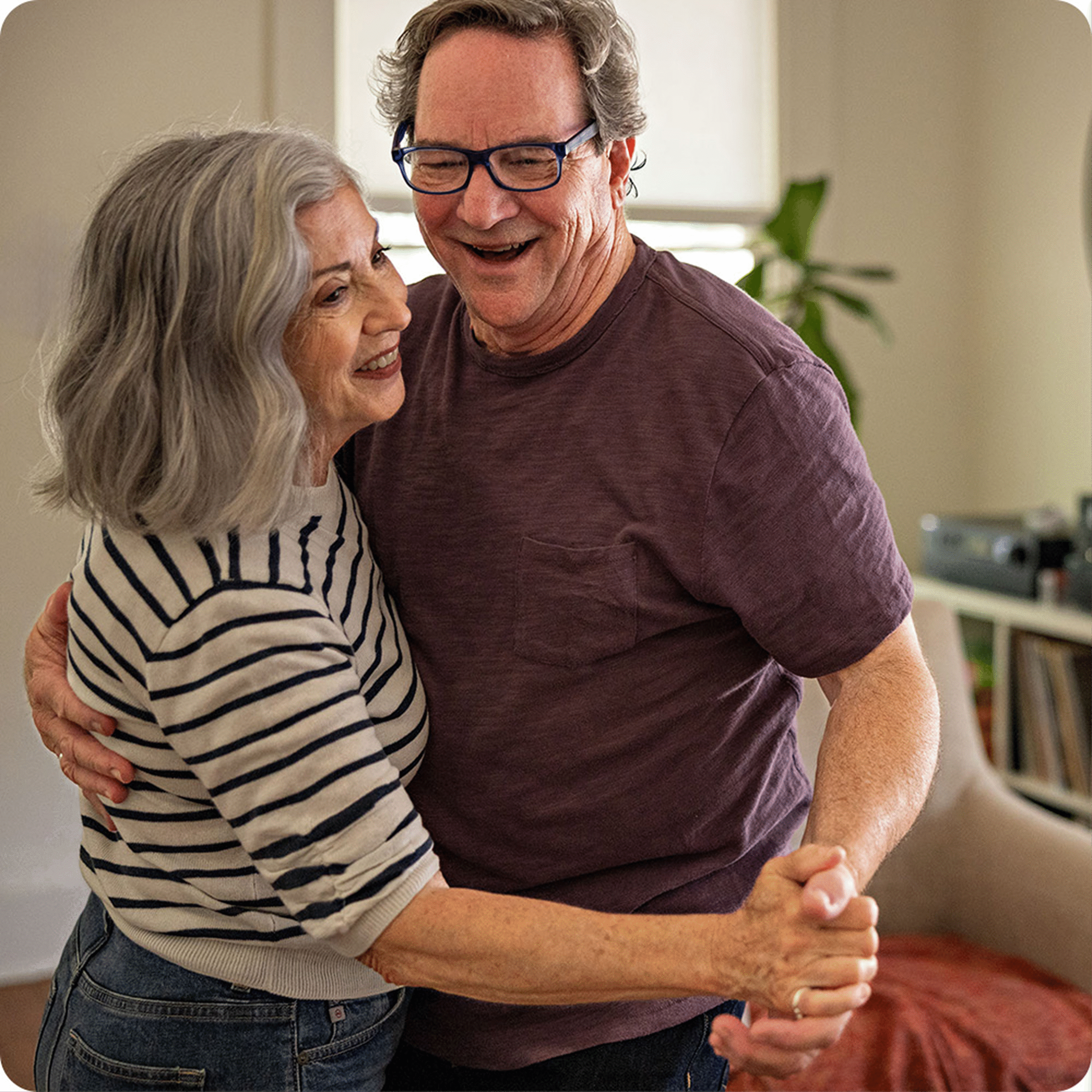 Older couple smiling and dancing closely indoors, with the woman wearing a striped shirt and the man in a purple shirt and glasses.
