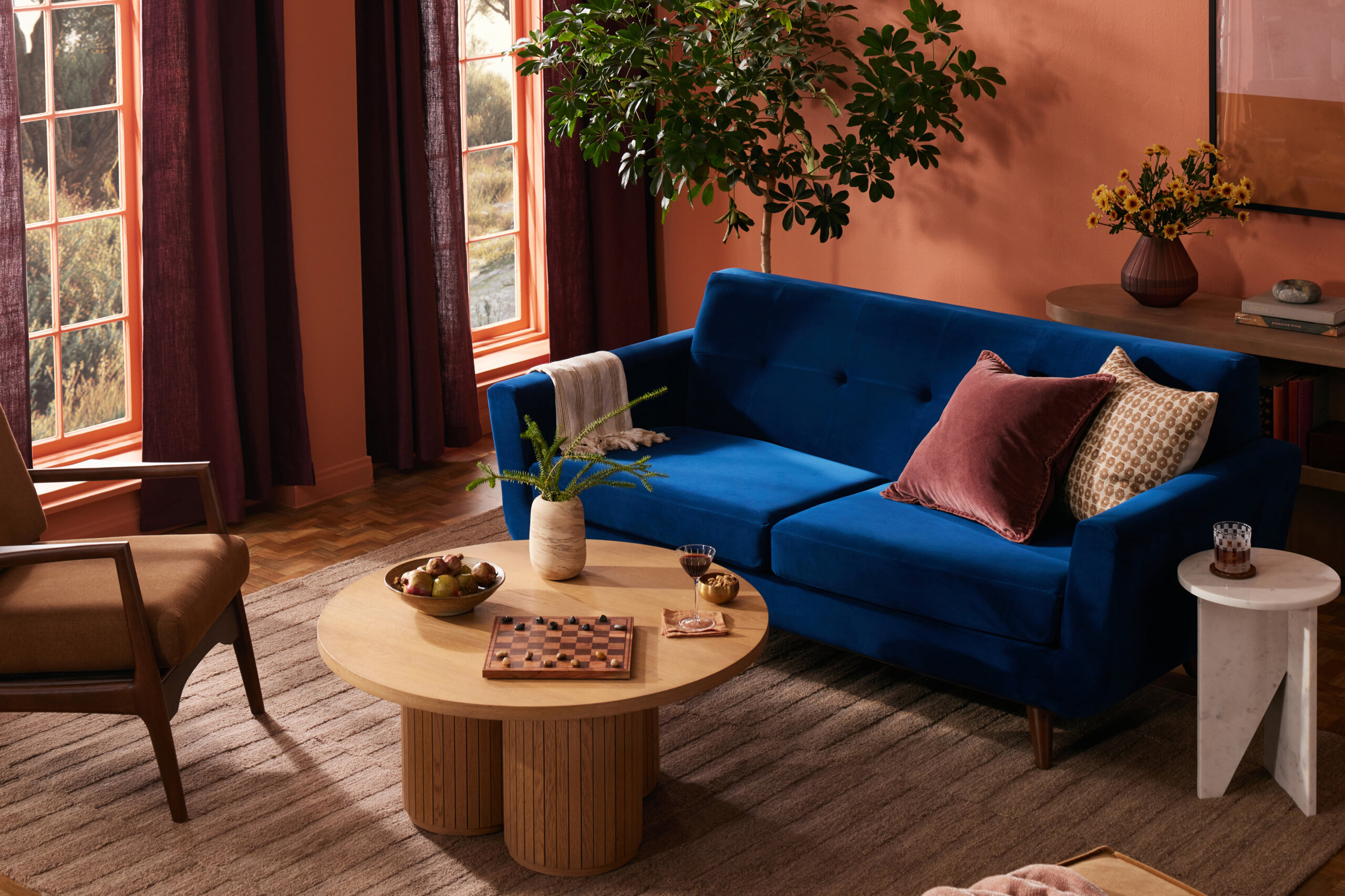 Cozy living room with a deep blue sofa adorned with two throw pillows and a beige blanket, a round wooden coffee table with a vase, bowl of fruit, wine glass, and checkerboard, large windows with maroon curtains, brown armchair, beige area rug, potted plant, and side tables with decor including a vase with flowers and books against a terracotta wall.
