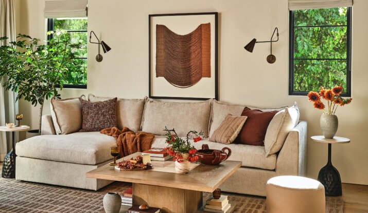 Cozy living room with a beige sectional sofa adorned with brown and beige cushions, a wooden coffee table with books, decorative vases, and fruits, two windows with greenery outside, and matching side tables with plants and an orange flower arrangement.
