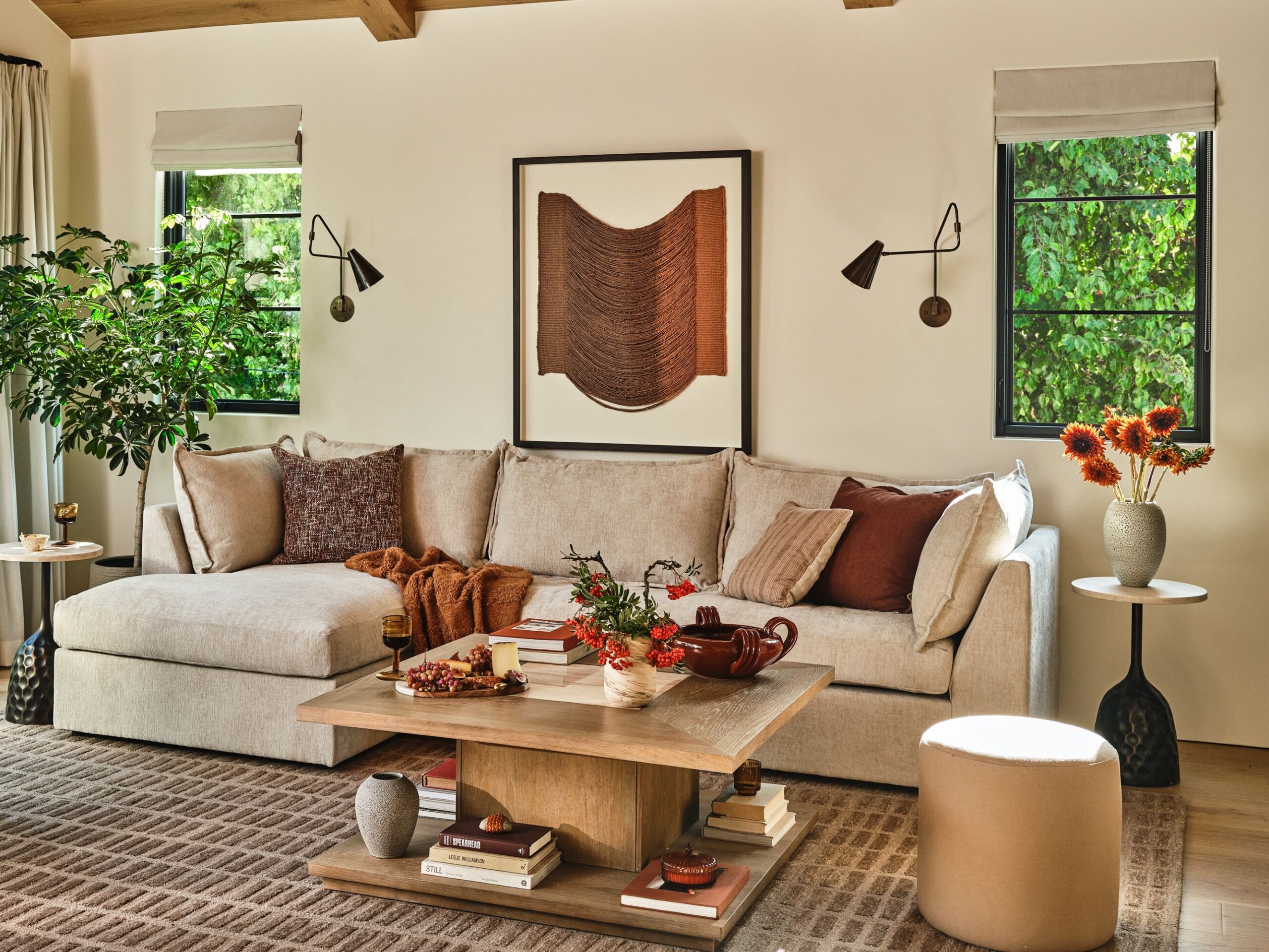 Cozy living room with a beige sectional sofa adorned with brown and beige cushions, a wooden coffee table with books, decorative vases, and fruits, two windows with greenery outside, and matching side tables with plants and an orange flower arrangement.