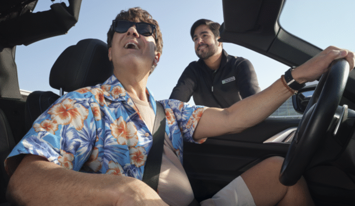 Man wearing a blue floral shirt and sunglasses sitting in the driver's seat of a convertible car, smiling and looking up, while another man leans in from outside the car, smiling back at him.