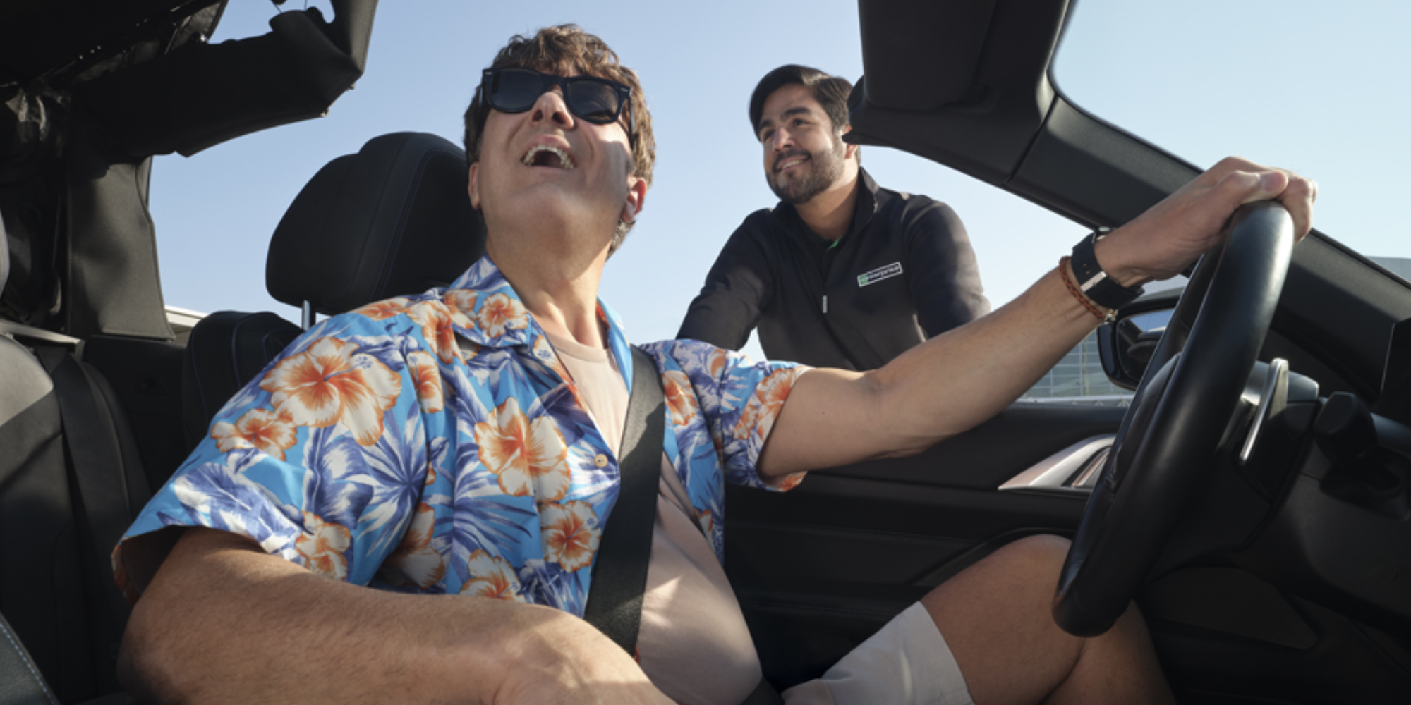 Man wearing a blue floral shirt and sunglasses sitting in the driver's seat of a convertible car, smiling and looking up, while another man leans in from outside the car, smiling back at him.