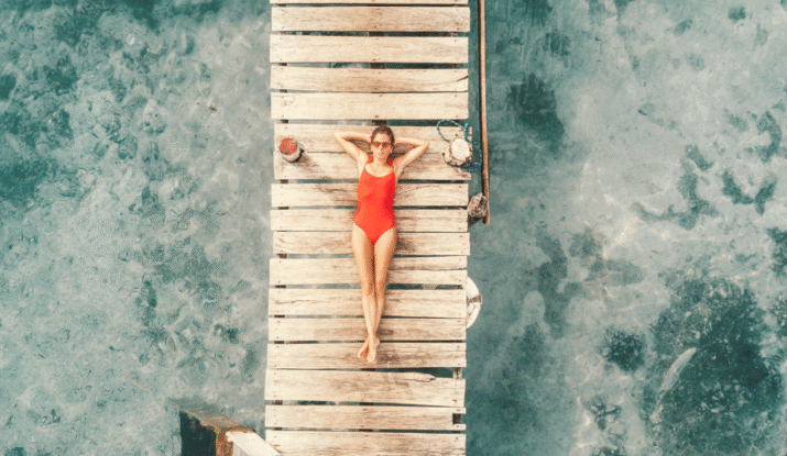 A woman in a red swimsuit and sunglasses lying on her back on a weathered wooden dock above clear blue water, with her arms resting behind her head and legs stretched out.