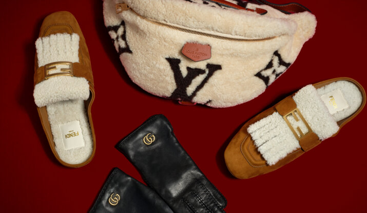 Pair of brown Fendi slip-on shoes with white shearling details and gold Fendi buckles, a cream Louis Vuitton shearling crossbody bag with a brown strap and large black LV logo, and a pair of black leather Gucci gloves with gold double G logos, all arranged on a dark red background.