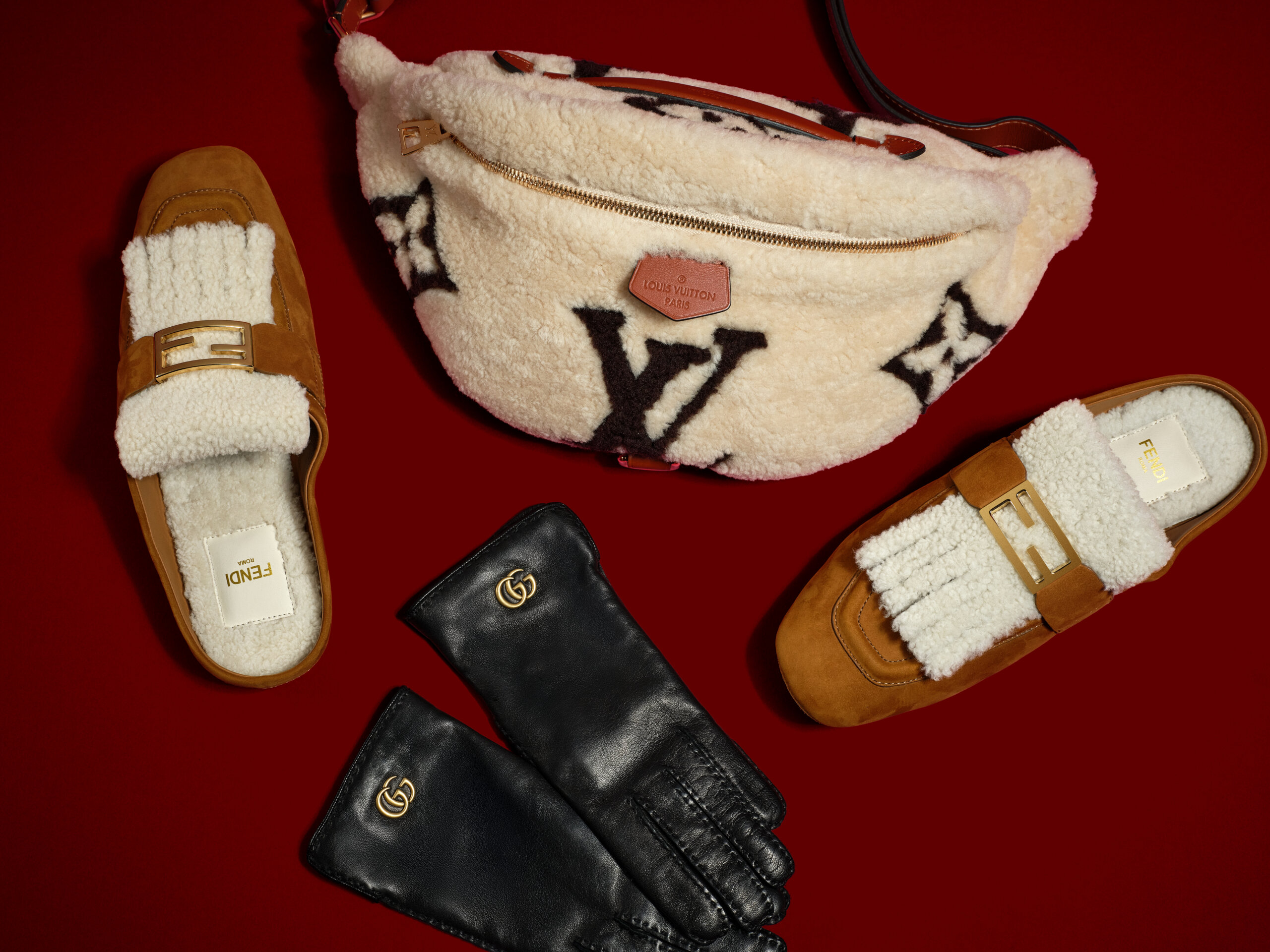 Pair of brown Fendi slip-on shoes with white shearling details and gold Fendi buckles, a cream Louis Vuitton shearling crossbody bag with a brown strap and large black LV logo, and a pair of black leather Gucci gloves with gold double G logos, all arranged on a dark red background.