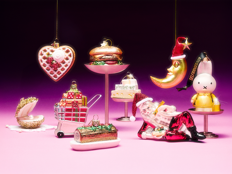 A collection of shiny Christmas ornaments on a pink surface with a dark purple background, including a heart-shaped waffle, a cheeseburger on a stand, a moon wearing a Santa hat, a white bunny figurine, a gold clam shell with a pearl, a shopping cart filled with gifts, a log cake, a white layered cake on a stand, and a reclining Santa Claus holding a gift box.