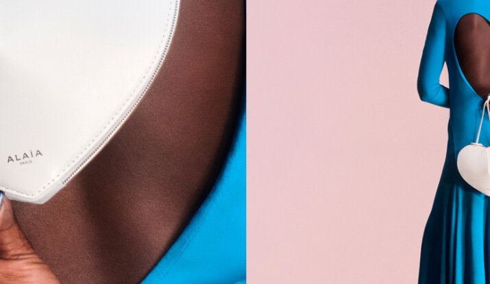Close-up of a person in a blue dress with a heart-shaped open back holding a white heart-shaped bag with the brand name Alaïa Paris on it against a pink background
