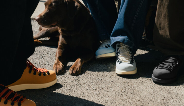 A brown dog lying on a paved surface next to three people wearing colorful shoes and jeans, with one person holding the dog's leash while sitting on a bench.