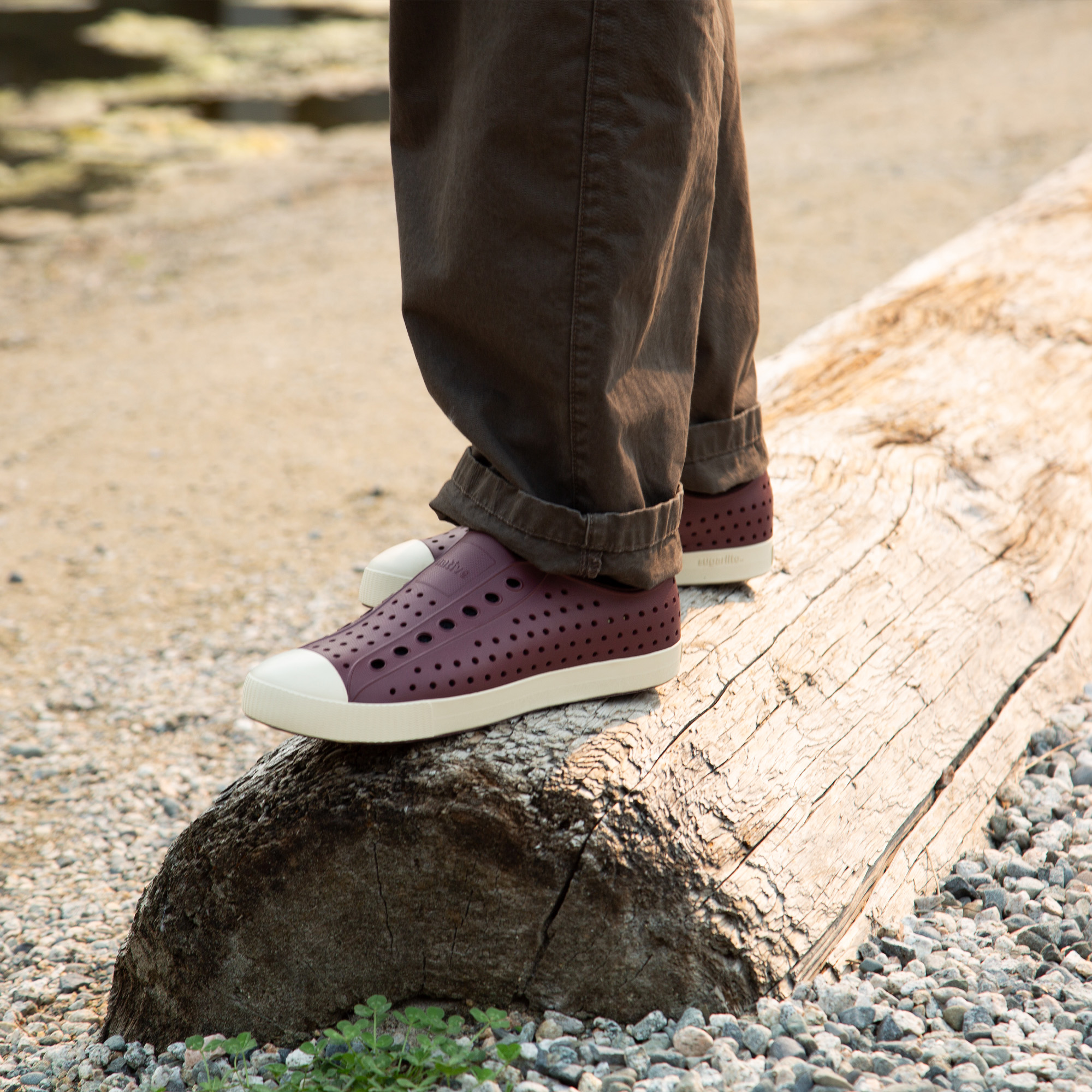 Person wearing brown pants and maroon slip-on shoes with white soles and toe caps standing on a large weathered log surrounded by gravel and small green plants.