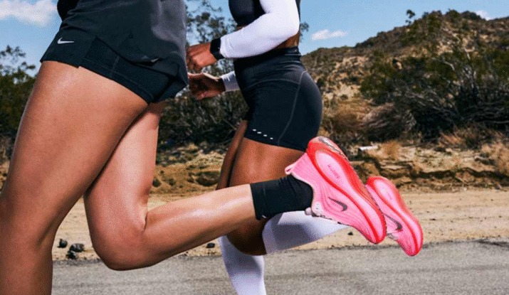 Close-up of two people running outdoors on a paved road with desert vegetation and hills in the background, both wearing athletic shorts and bright pink Nike running shoes.