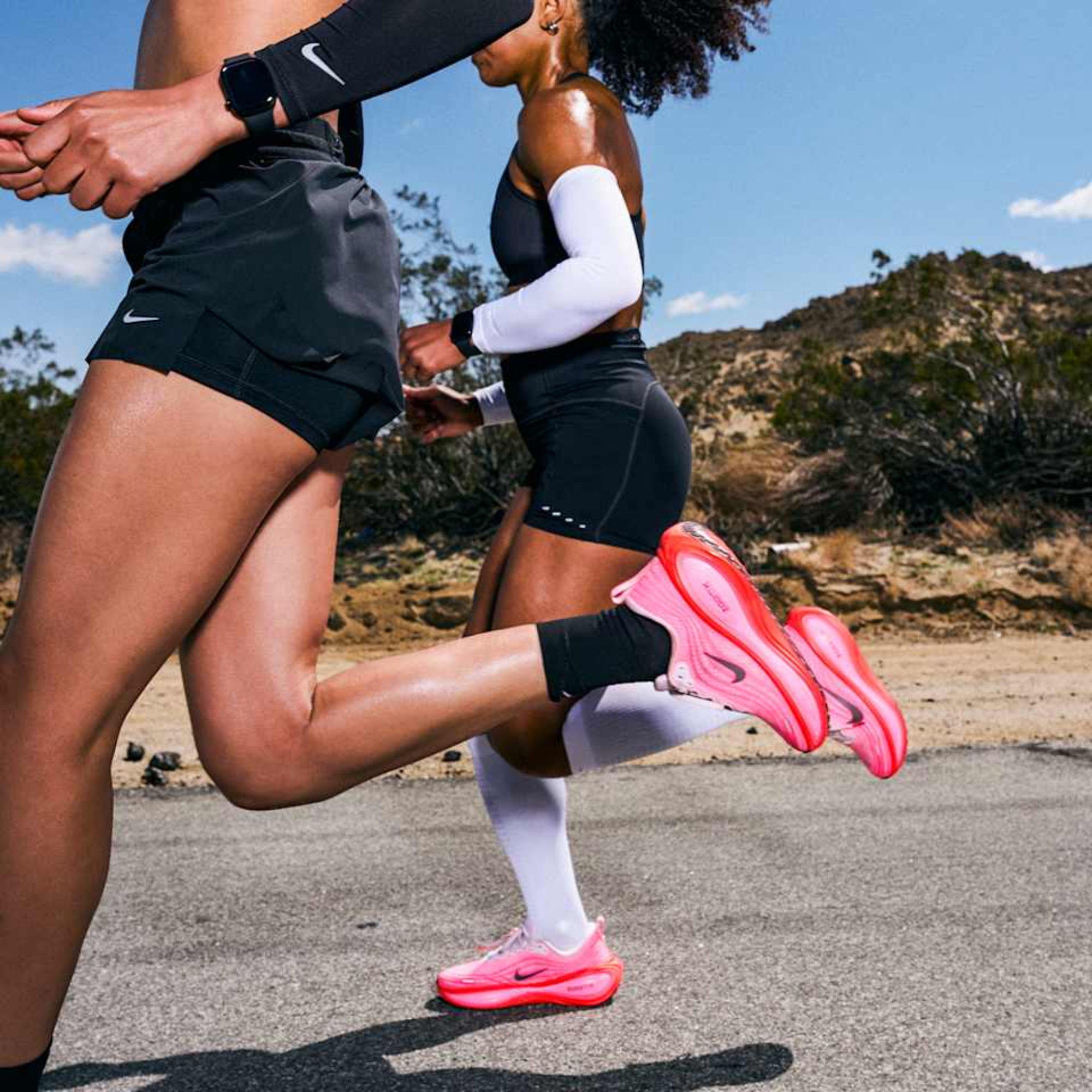 Close-up of two people running outdoors on a paved road with desert vegetation and hills in the background, both wearing athletic shorts and bright pink Nike running shoes.