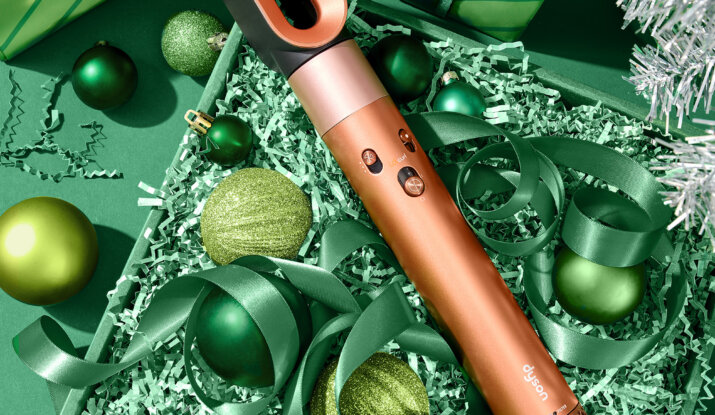 Copper-colored Dyson hair styling tool surrounded by green ribbon and green Christmas ornaments in a green gift box with additional wrapped gifts and a silver tinsel tree branch in the background.