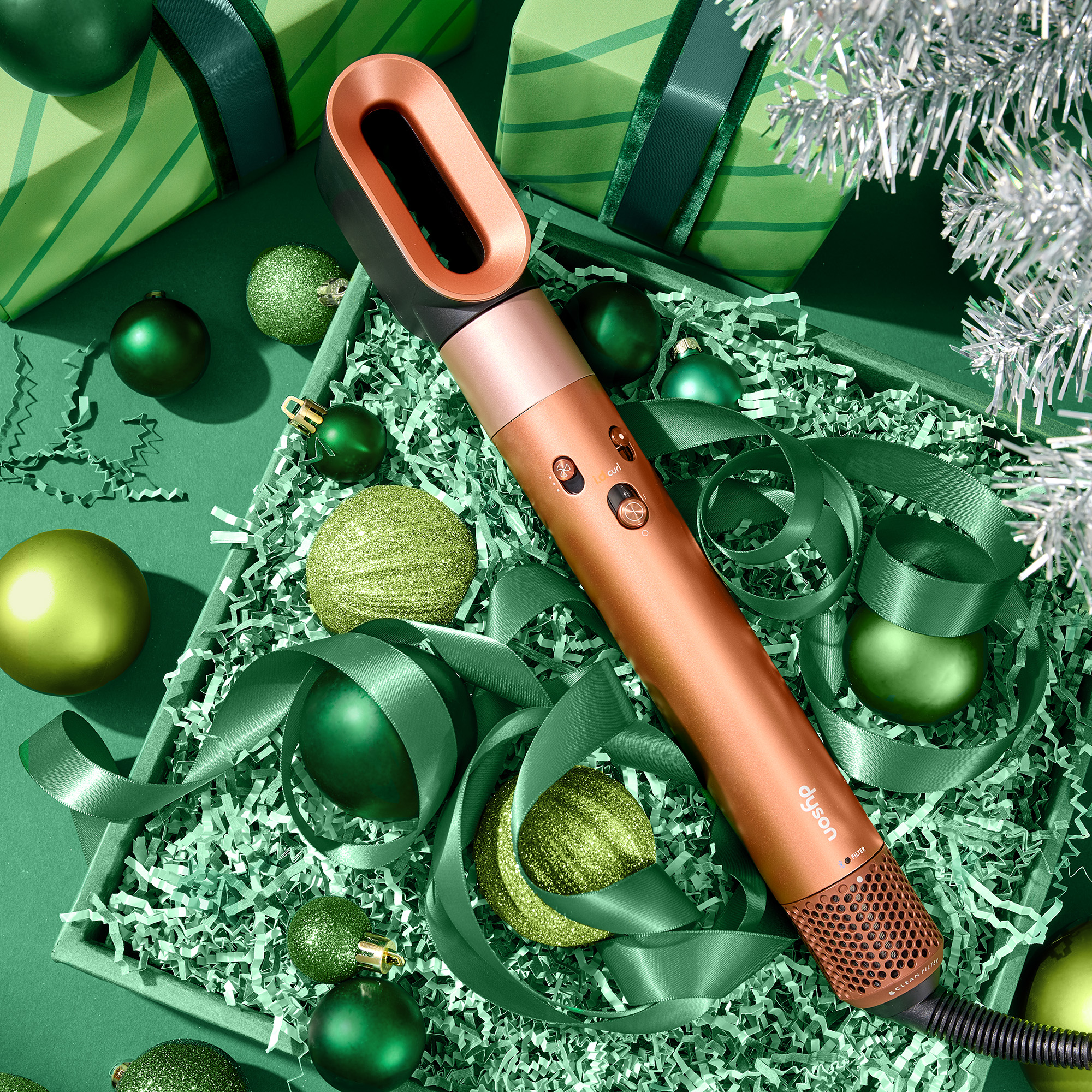 Copper-colored Dyson hair styling tool surrounded by green ribbon and green Christmas ornaments in a green gift box with additional wrapped gifts and a silver tinsel tree branch in the background.