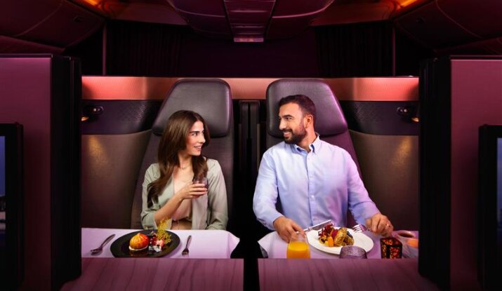 A man and a woman seated in a business class airplane cabin, enjoying meals and looking at each other while smiling, with ambient red lighting.