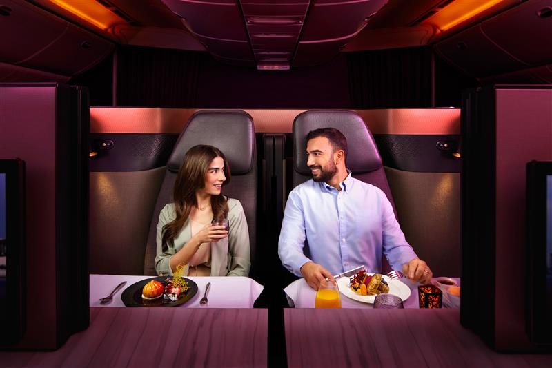 A man and a woman seated in a business class airplane cabin, enjoying meals and looking at each other while smiling, with ambient red lighting.
