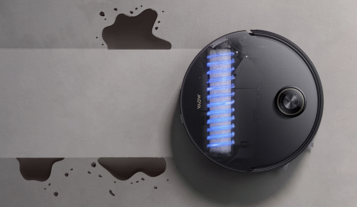 Top view of a black robotic vacuum cleaner cleaning a dark liquid spill on a light grey floor, with a highlighted blue cylindrical brush visible through a transparent section on the vacuum's top surface.