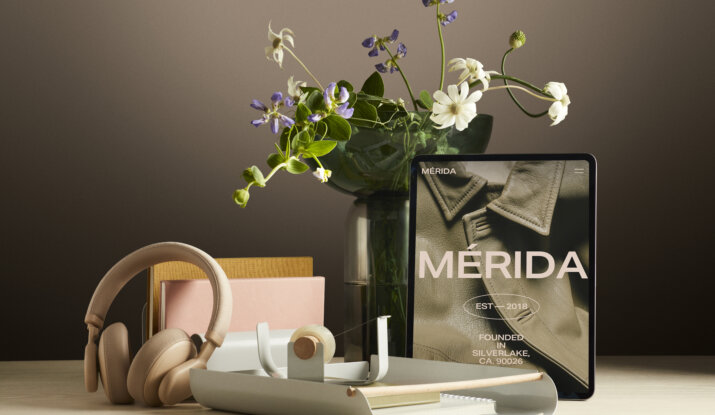 Desk setup with beige wireless headphones, notebooks in yellow and pink, a tape dispenser, and a vase with green and white flowers behind a tablet displaying a close-up of a leather jacket with the text 'MÉRIDA EST 2018 FOUNDED IN SILVERLAKE, CA. 90026'