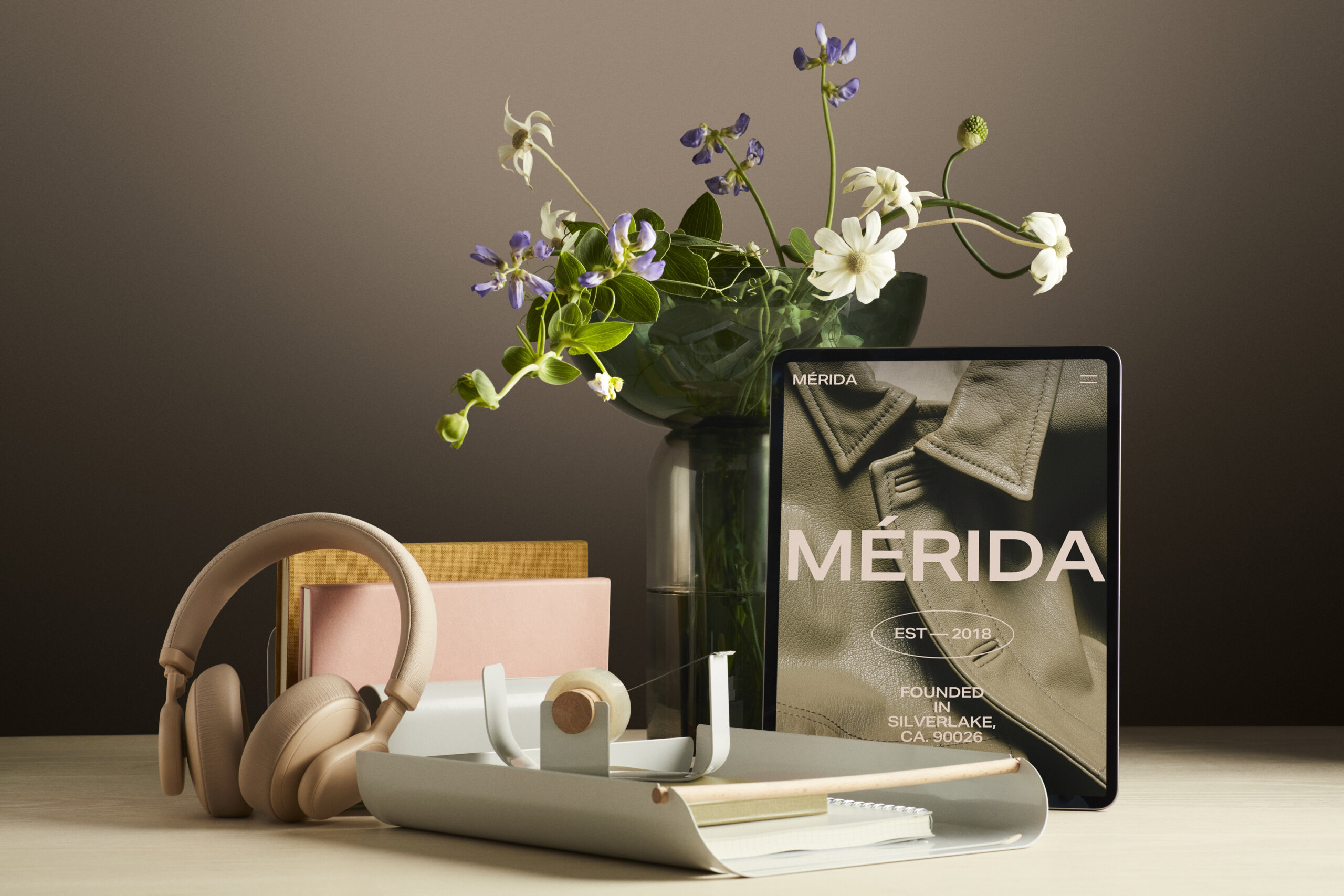 Desk setup with beige wireless headphones, notebooks in yellow and pink, a tape dispenser, and a vase with green and white flowers behind a tablet displaying a close-up of a leather jacket with the text 'MÉRIDA EST 2018 FOUNDED IN SILVERLAKE, CA. 90026'