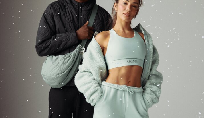 A man and a woman posing together against a neutral background with white specks; the man is wearing sunglasses, a black quilted jacket, black pants, and a light green quilted sling bag, while the woman is dressed in a light green sports bra, matching sweatpants, and a fuzzy jacket draped over her shoulders with her hands in her pockets.