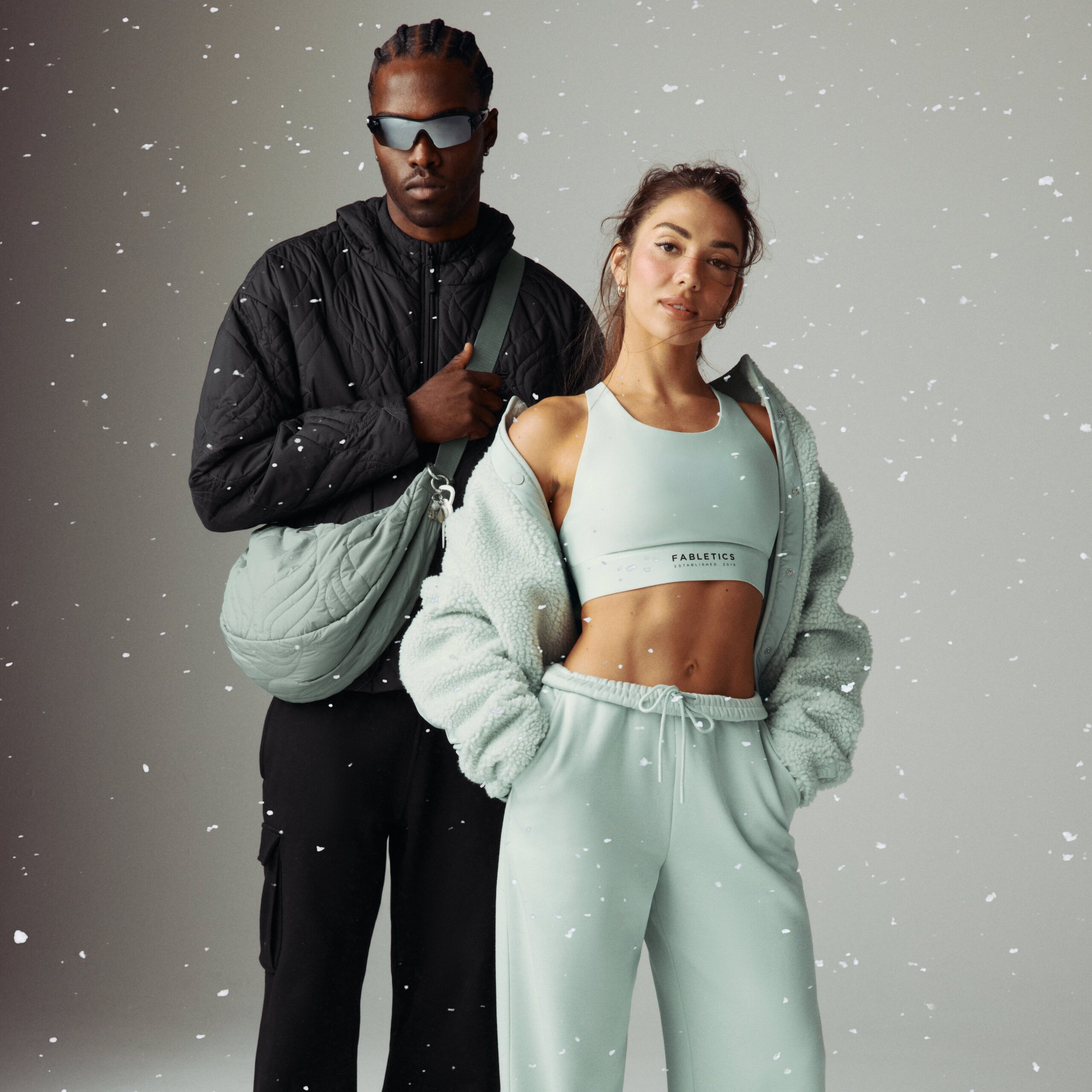 A man and a woman posing together against a neutral background with white specks; the man is wearing sunglasses, a black quilted jacket, black pants, and a light green quilted sling bag, while the woman is dressed in a light green sports bra, matching sweatpants, and a fuzzy jacket draped over her shoulders with her hands in her pockets.
