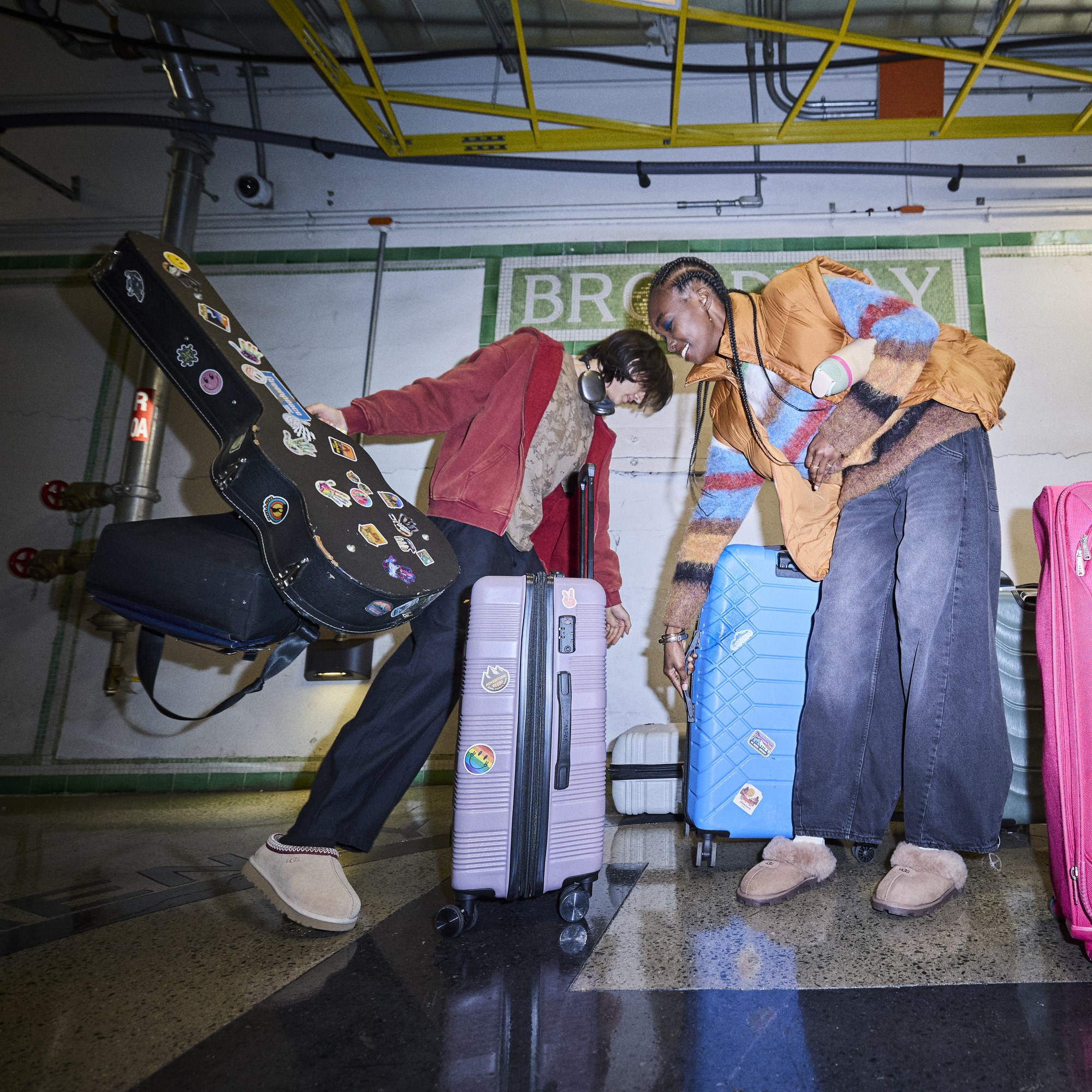 Two people standing indoors with colorful suitcases and a guitar case covered in stickers, one person bending forward while the other reaches down toward a blue suitcase, in front of a tiled wall with partially visible text.