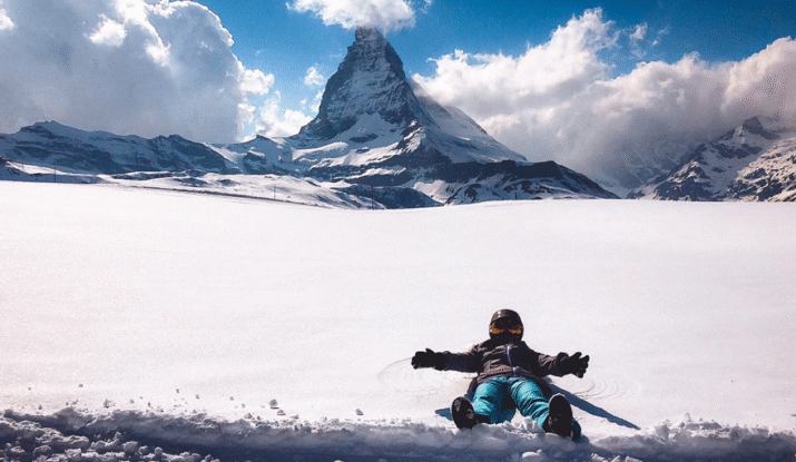 Person lying on snow with arms outstretched in front of the Matterhorn mountain covered partially by clouds under a blue sky.