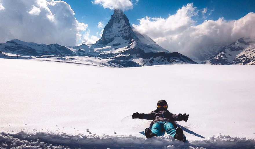 Person lying on snow with arms outstretched in front of the Matterhorn mountain covered partially by clouds under a blue sky.