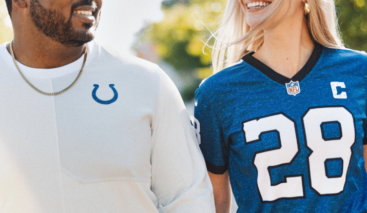 A smiling man and woman walking outdoors, the man wearing glasses, a gold chain, and a white shirt with a blue horseshoe logo, and the woman wearing a blue NFL jersey with the number 28 and a captain's patch, both looking at each other against a blurred background of trees and sunlight.