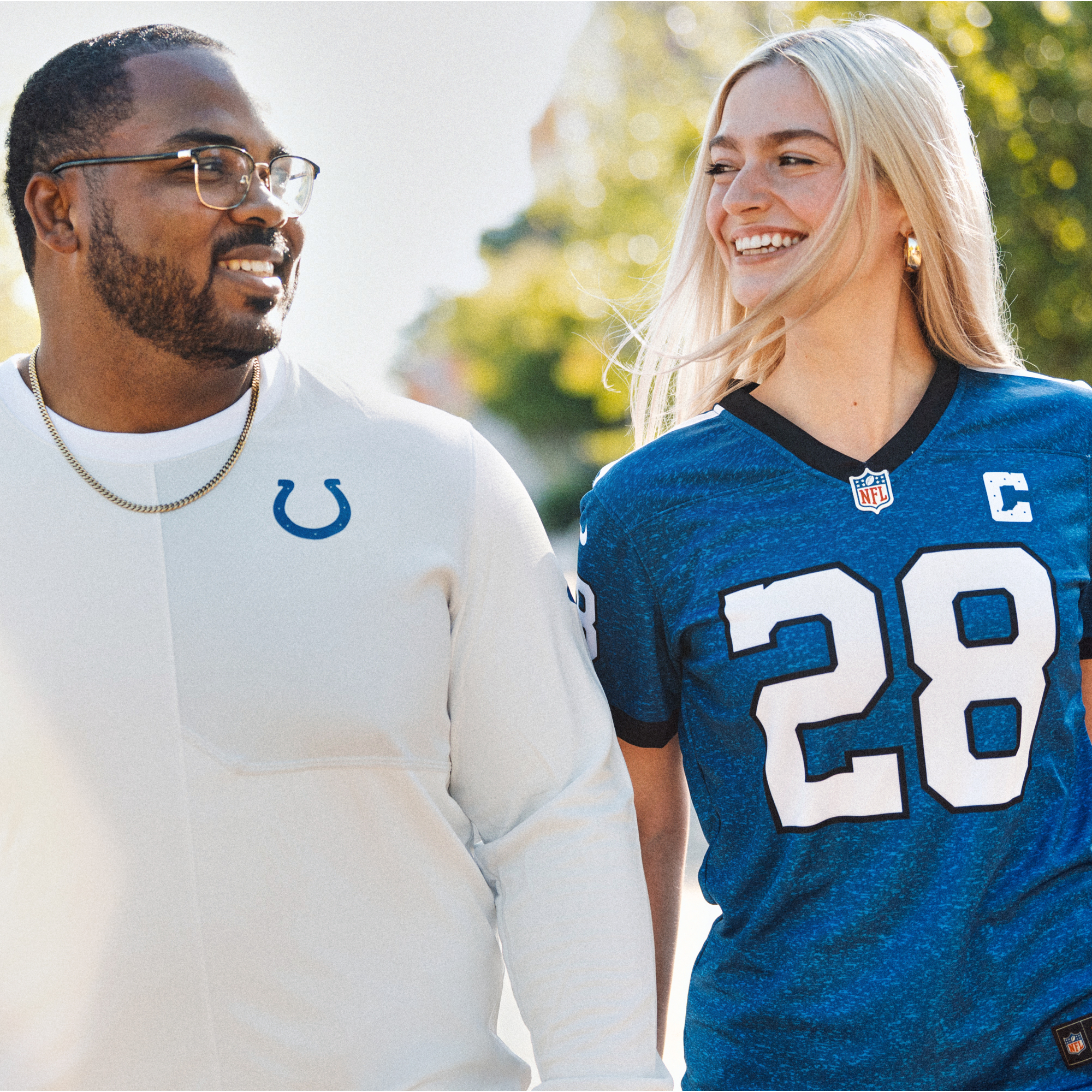 A smiling man and woman walking outdoors, the man wearing glasses, a gold chain, and a white shirt with a blue horseshoe logo, and the woman wearing a blue NFL jersey with the number 28 and a captain's patch, both looking at each other against a blurred background of trees and sunlight.