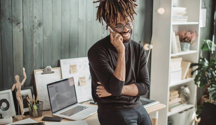 Smiling man with dreadlocks and glasses standing in a modern office, talking on a smartphone, with a laptop and various office supplies on the desk behind him.