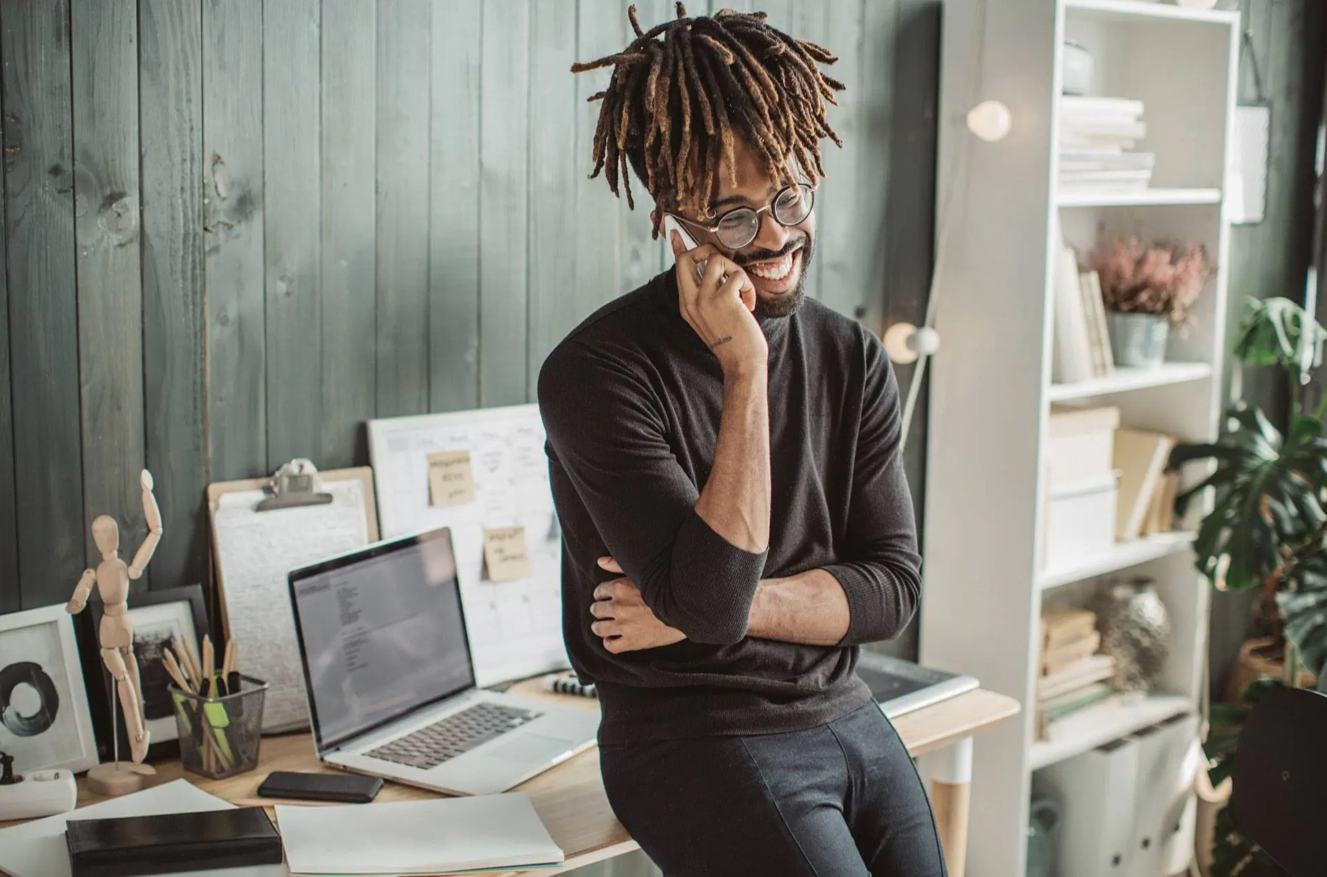 Smiling man with dreadlocks and glasses standing in a modern office, talking on a smartphone, with a laptop and various office supplies on the desk behind him.