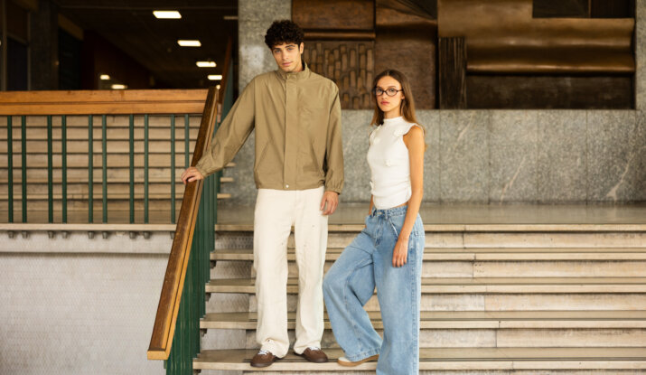 A young man in a beige jacket and white pants leaning against a stair railing next to a young woman wearing glasses, a white sleeveless top, and wide-leg blue jeans standing on marble stairs in an indoor setting with wooden and stone architectural details.