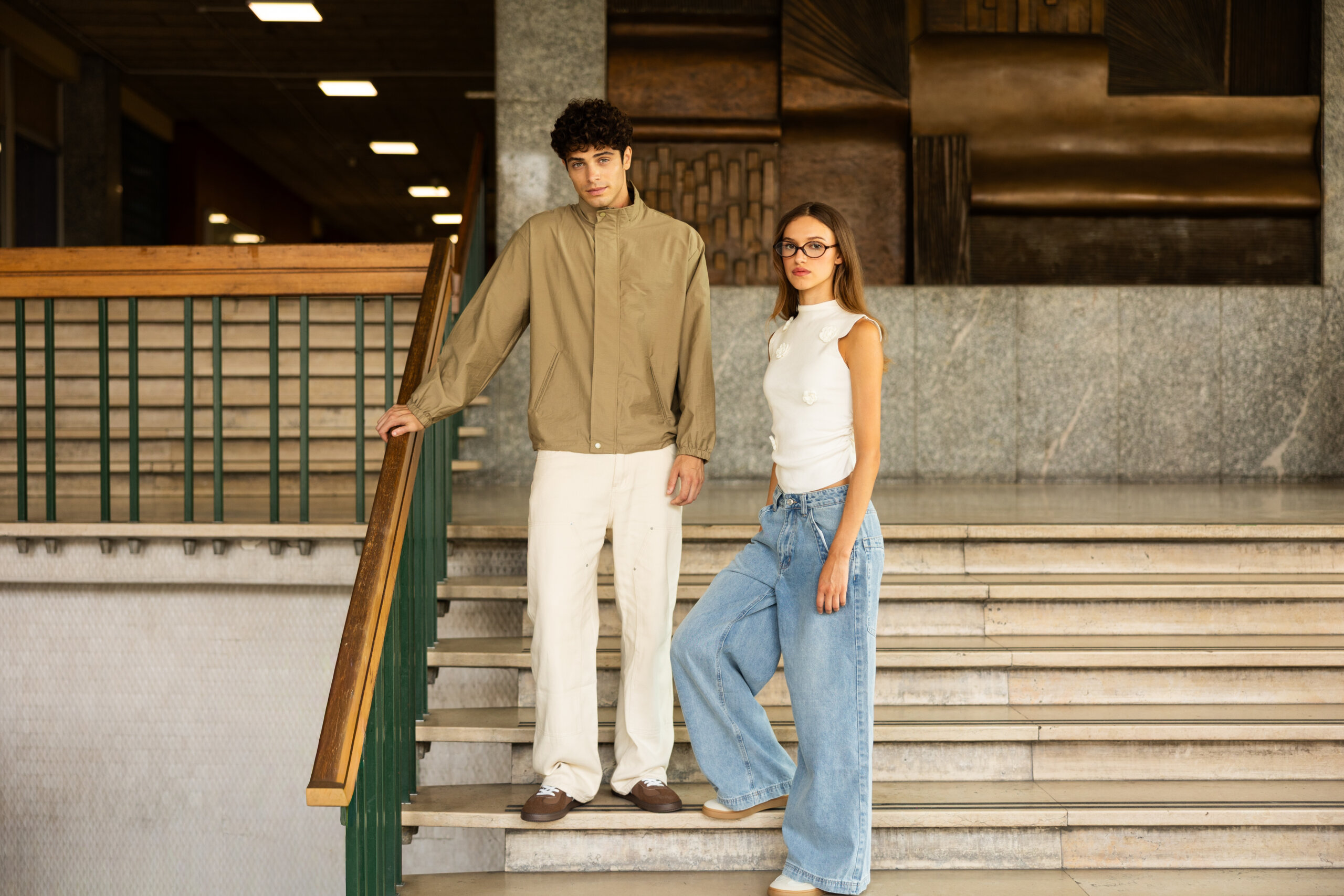 A young man in a beige jacket and white pants leaning against a stair railing next to a young woman wearing glasses, a white sleeveless top, and wide-leg blue jeans standing on marble stairs in an indoor setting with wooden and stone architectural details.