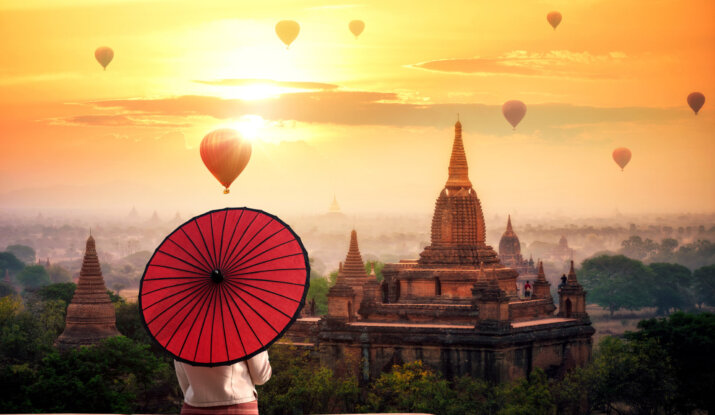 Person holding a red umbrella overlooking ancient temples at sunrise with hot air balloons floating in the sky
