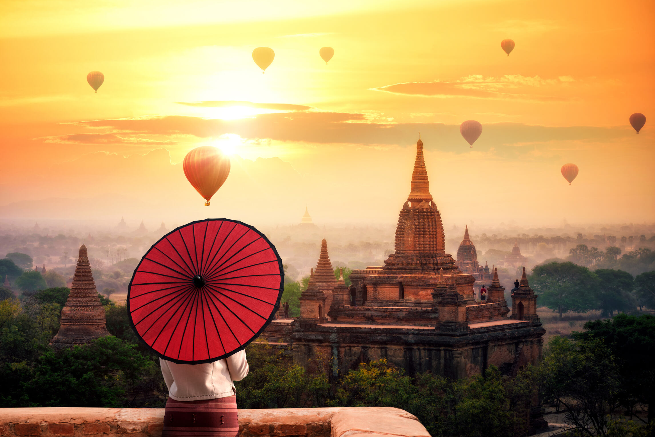 Person holding a red umbrella overlooking ancient temples at sunrise with hot air balloons floating in the sky