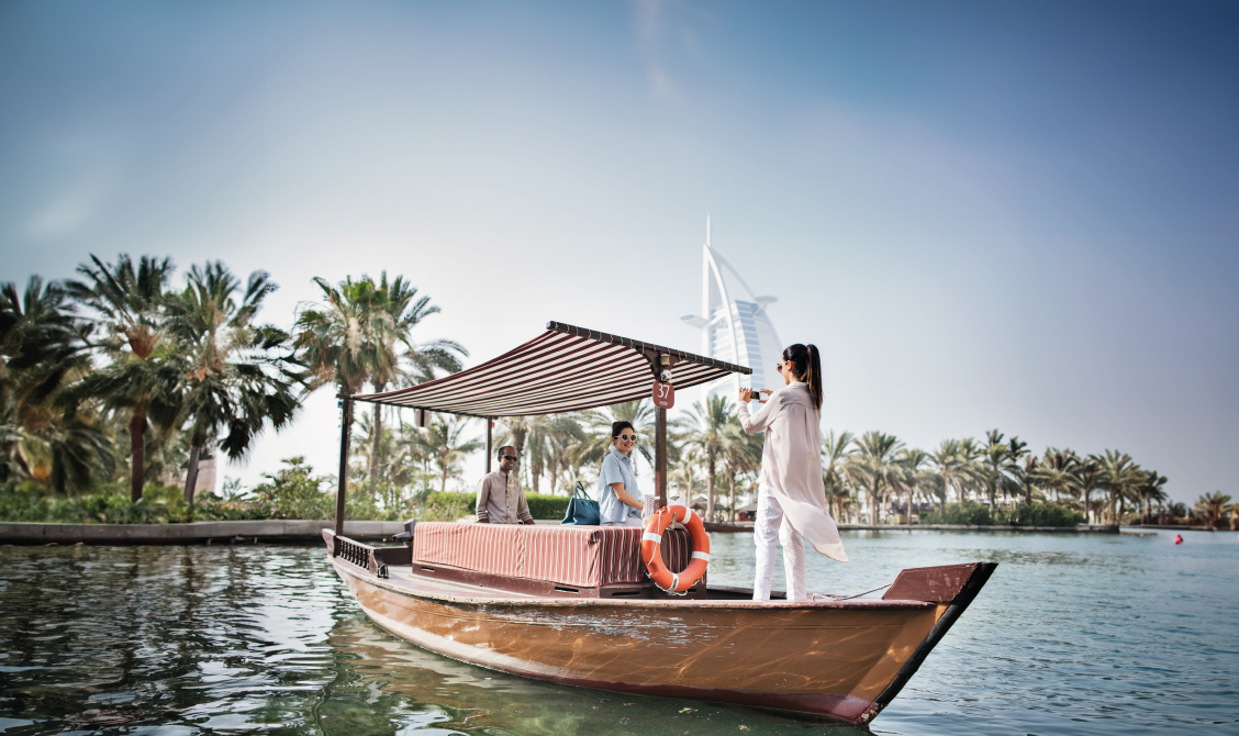 Two people sitting on a wooden boat with a striped canopy on a calm body of water, while a third person stands at the front of the boat taking a photo, with palm trees and the Burj Al Arab building visible in the background under a clear blue sky.