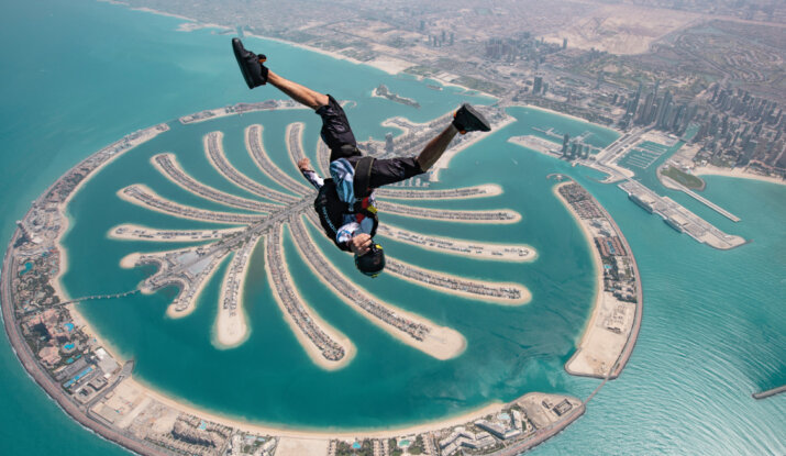 Skydiver in mid-air above the Palm Jumeirah island in Dubai, with the city's coastline and buildings visible in the background.