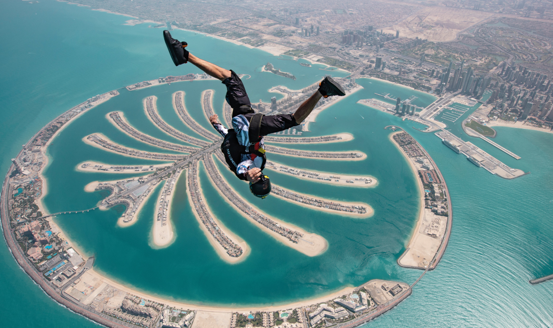Skydiver in mid-air above the Palm Jumeirah island in Dubai, with the city's coastline and buildings visible in the background.