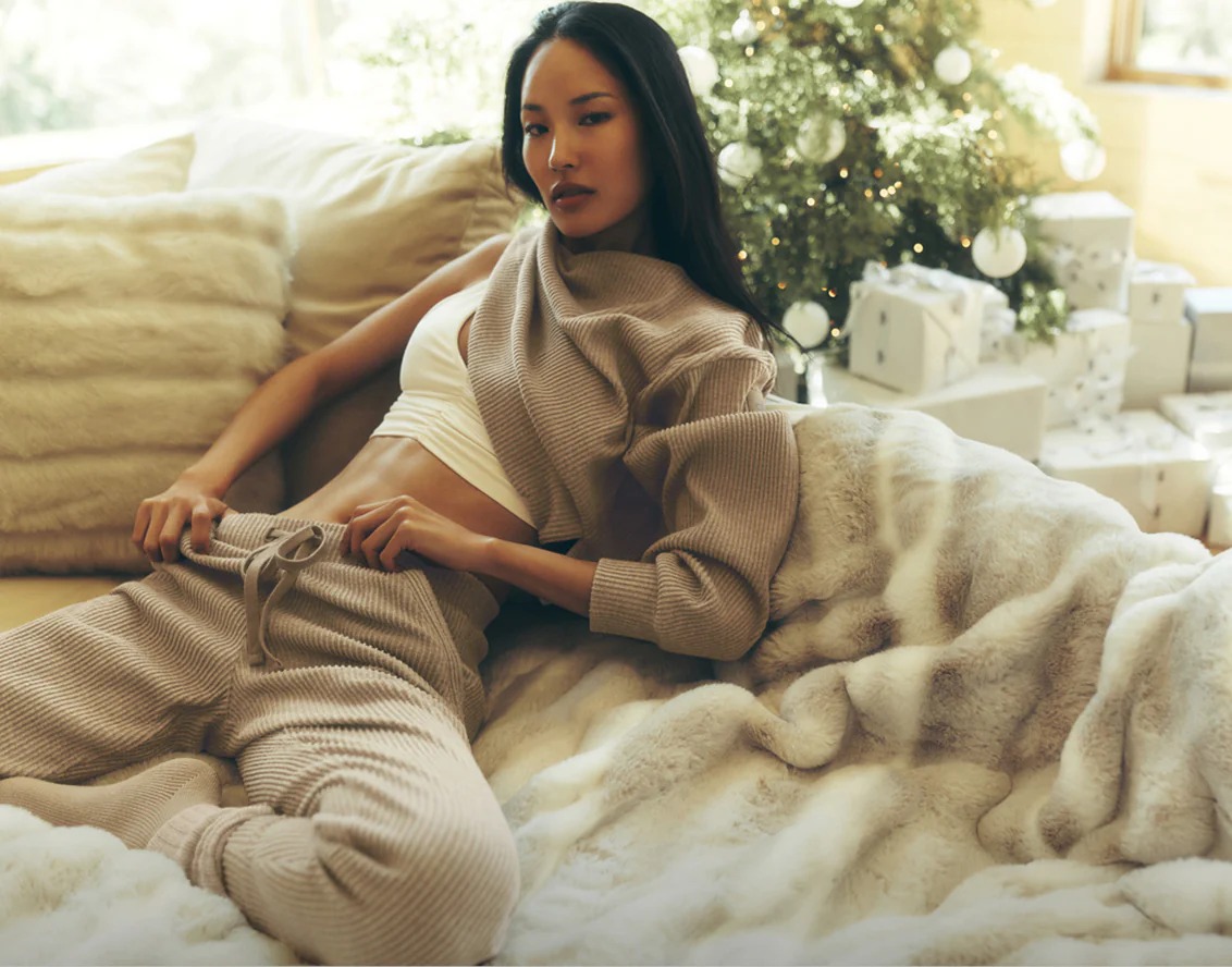 Young woman lounging on a plush, beige blanket with cream-colored cushions behind her and a decorated Christmas tree with wrapped gifts in the background, wearing beige knit pants and a cropped white top with a matching beige knit sweater draped over her shoulder.