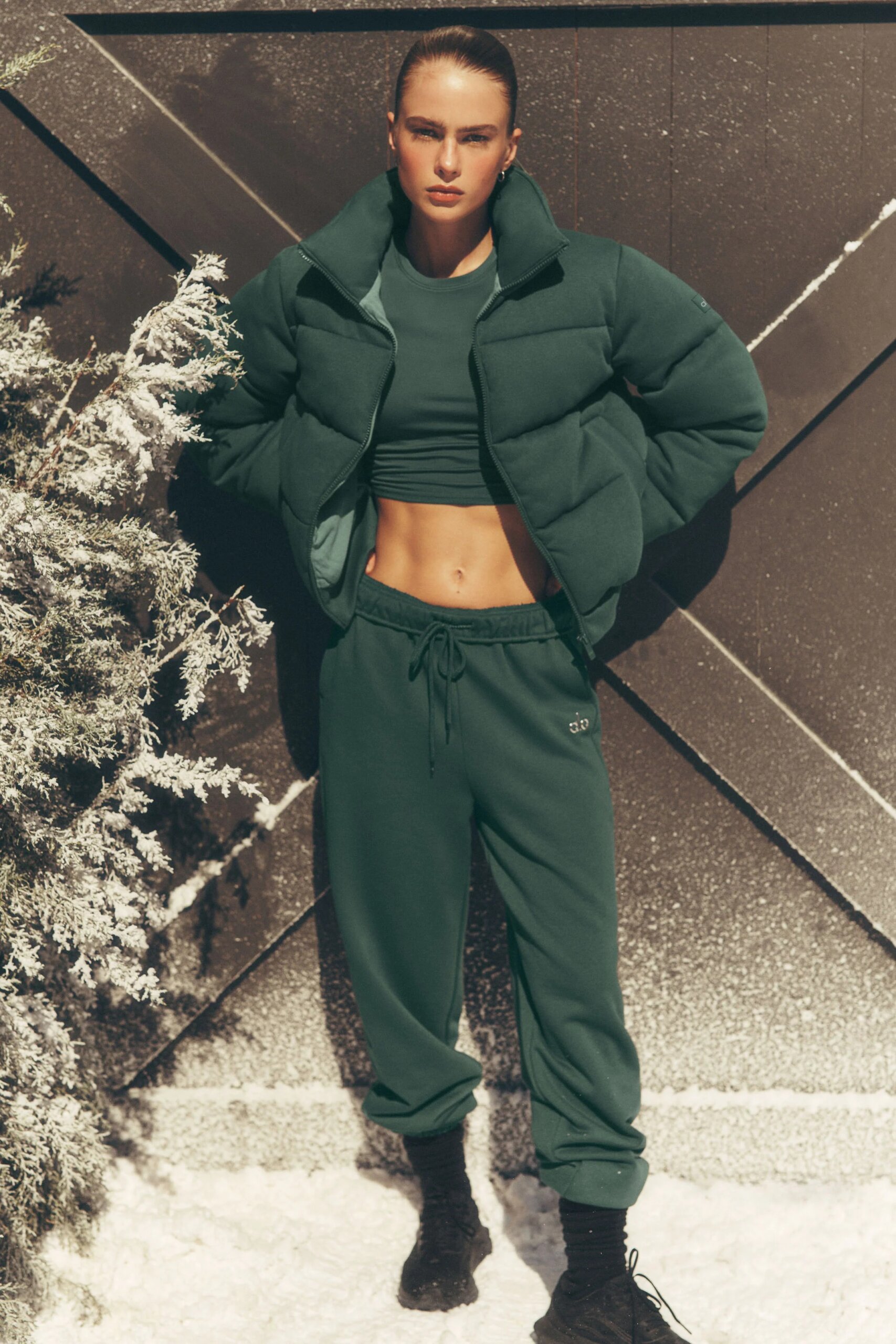 A person standing outdoors against a geometric black wall with snow on the ground, wearing a dark green puffer jacket, matching crop top, jogger pants, black socks, and black shoes, with their hands behind their back and a serious expression.