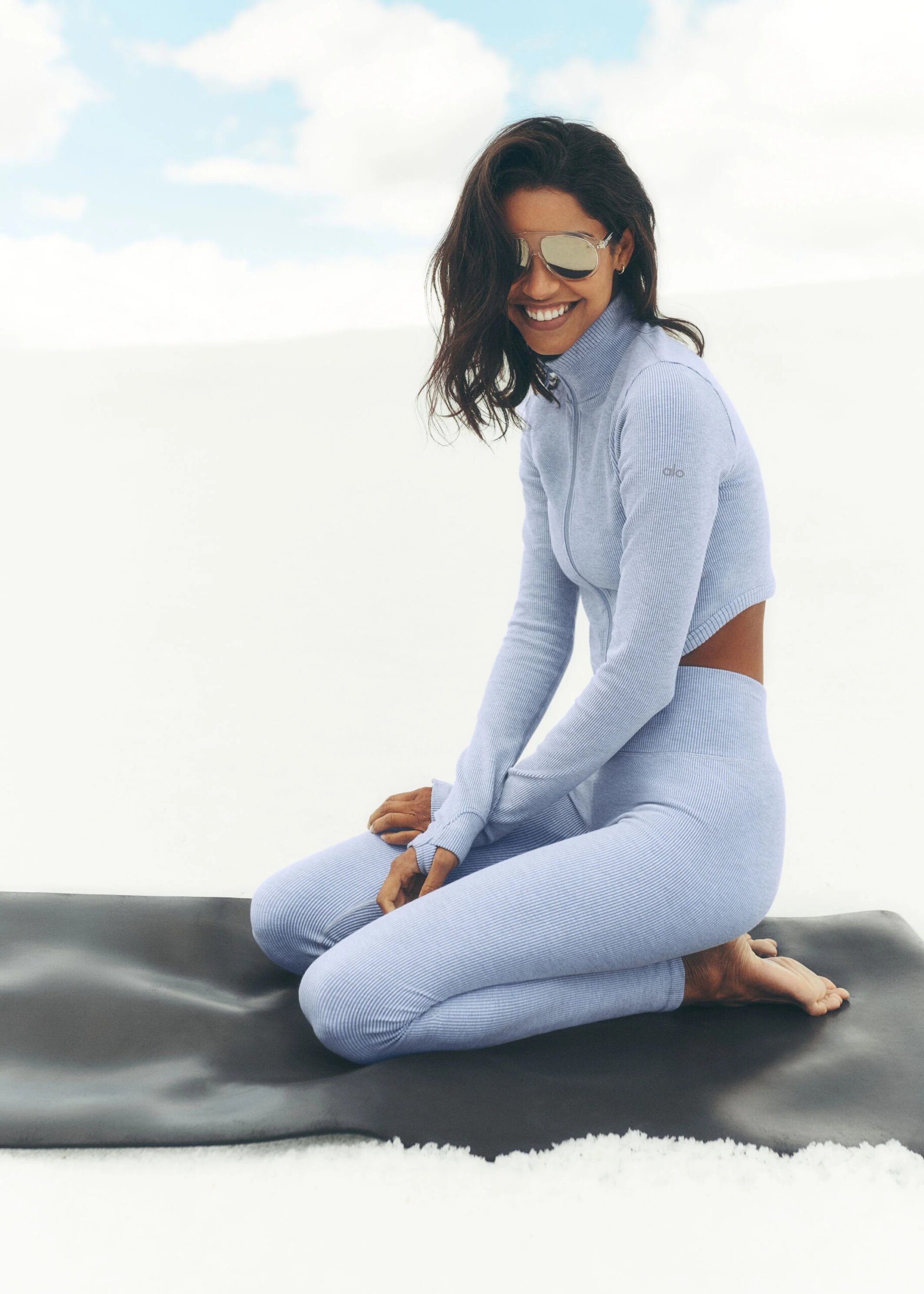 Smiling woman with long dark hair wearing reflective sunglasses and a light blue ribbed athletic outfit, kneeling on a black exercise mat against a white snowy background under a partly cloudy sky.