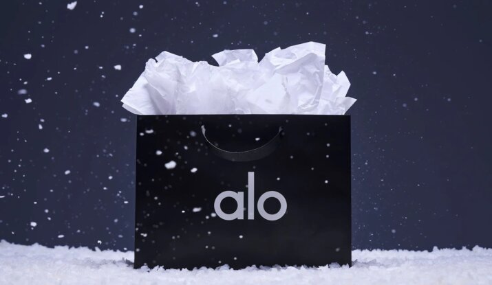 Black shopping bag with white tissue paper inside and the word 'alo' printed on it, surrounded by falling snow on a snowy surface against a dark background