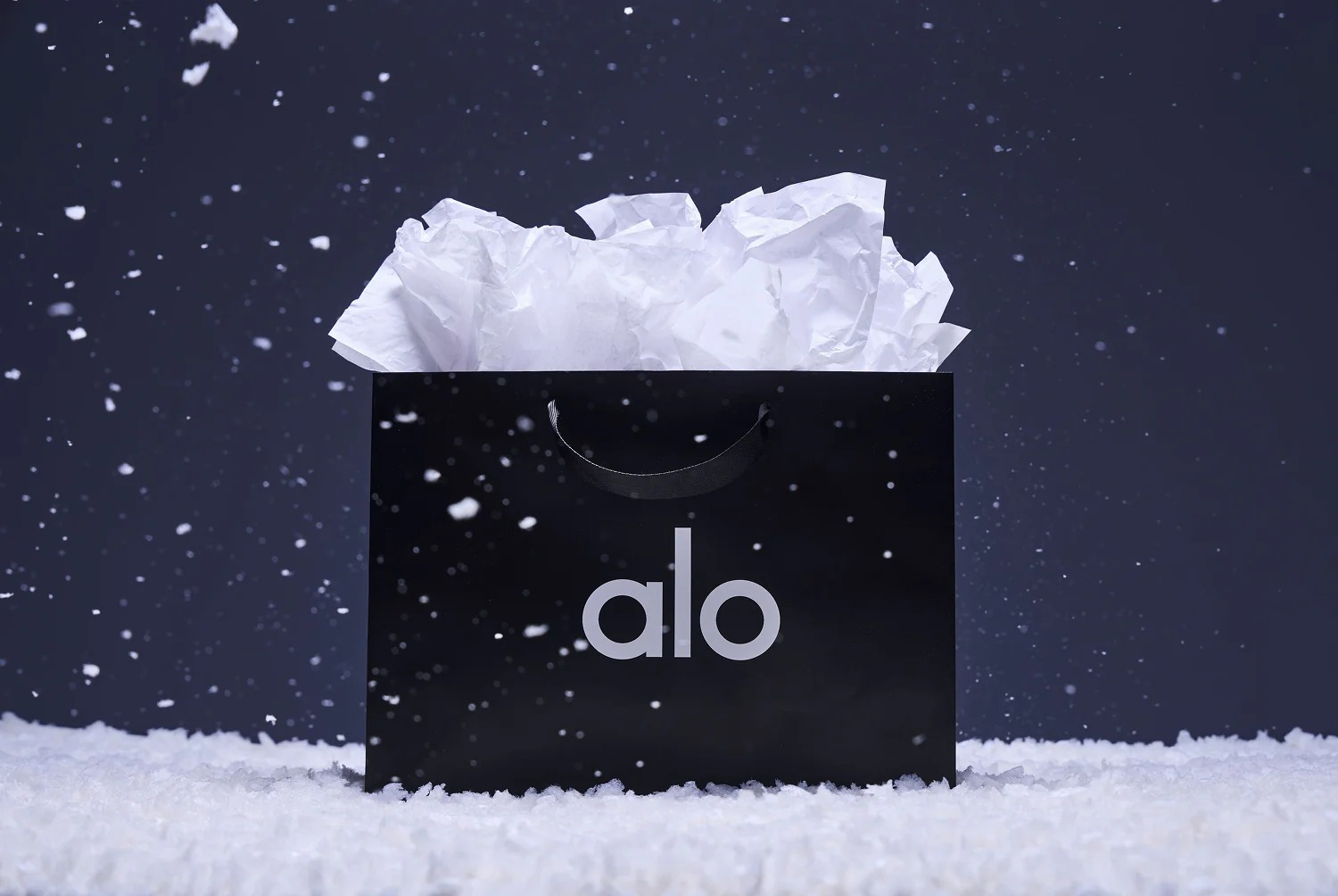 Black shopping bag with white tissue paper inside and the word 'alo' printed on it, surrounded by falling snow on a snowy surface against a dark background
