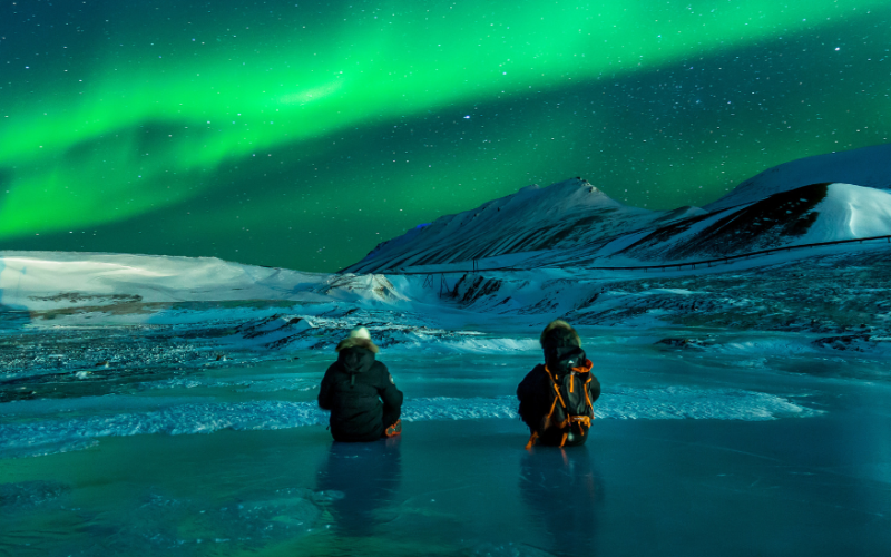 Two people watching northern lights over snowy mountains
